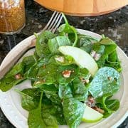 Spinach salad with maple dijon dressing and candied pecans on a white plate next to the serving bowl.